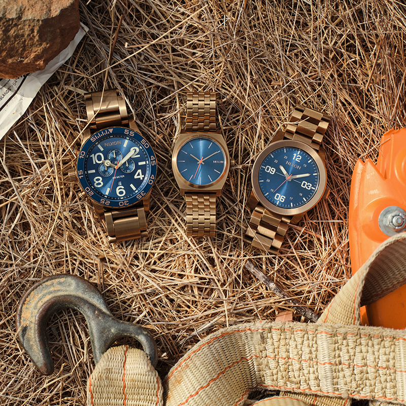 The 51-30 Chrono, Time Teller, and Corporal in the Desert Gold colorway, laying on top of pine needles with camping gear nearby.