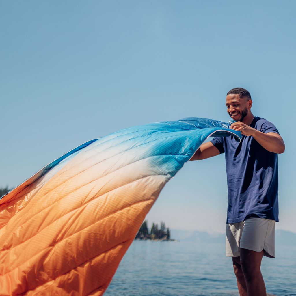 A man dusting out his Original Puffy Blanket in Sunset Fade before setting it down to enjoy the view by a lake.