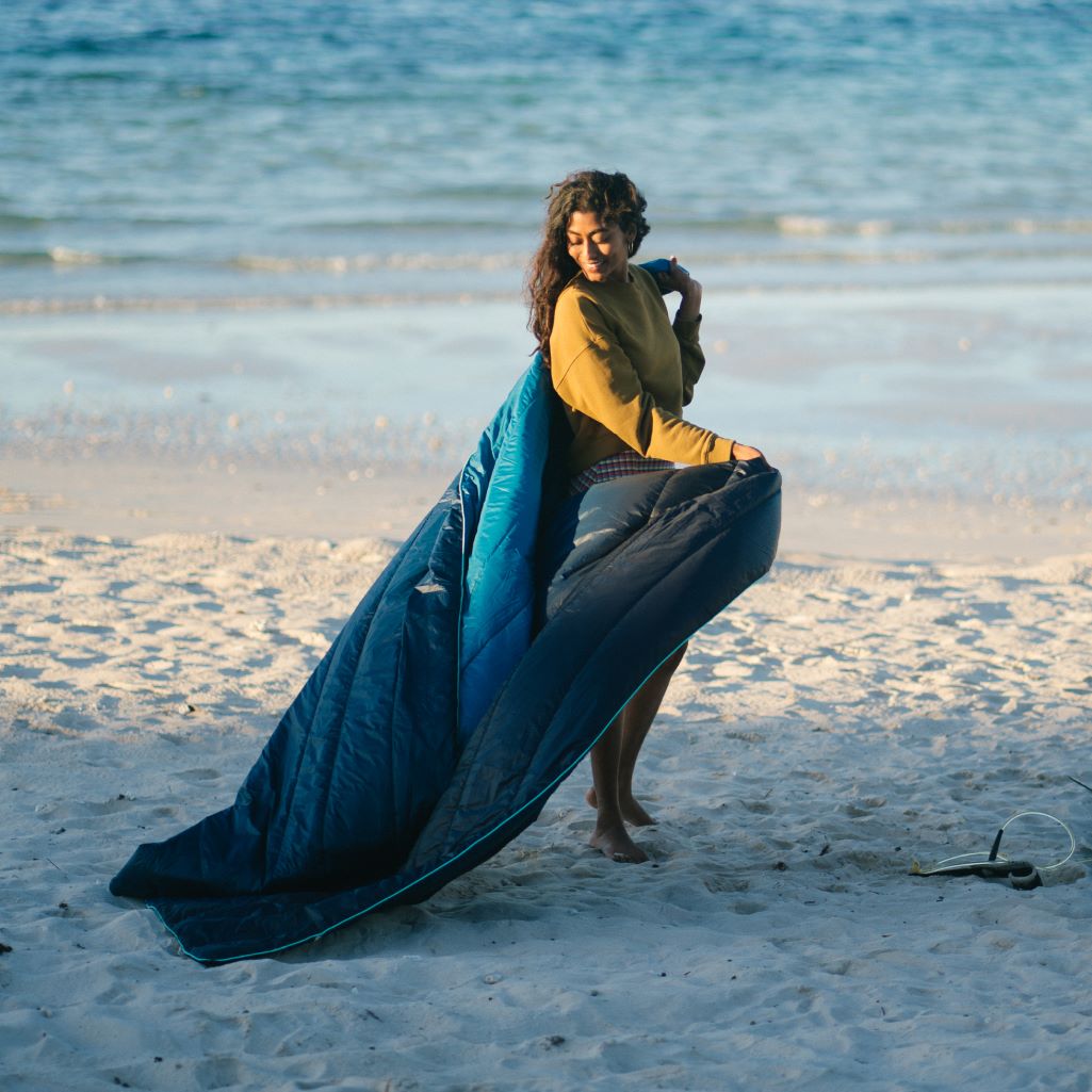 A person carries their 2-person sized Ocean Fade Rumpl blanket across the sand.