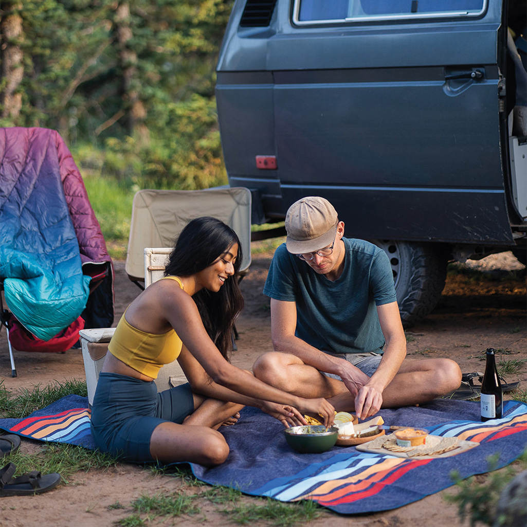 A couple uses the Rumpl Everywhere Mat as a waterproof picnic mat as they share a meal right outside their van in the great outdoors.