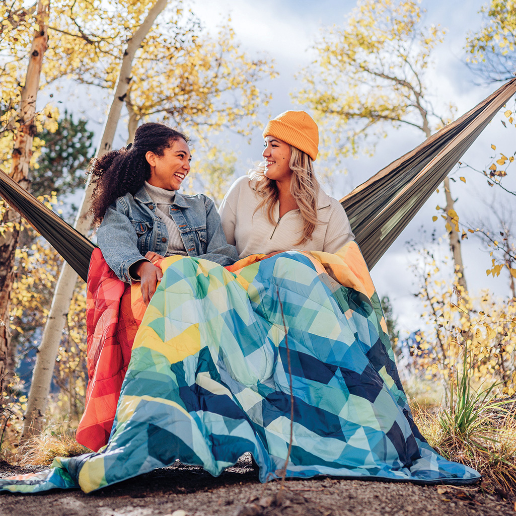 two friends sitting on a hammock and sharing a Rumpl Original Puffy Blanket to stay warm