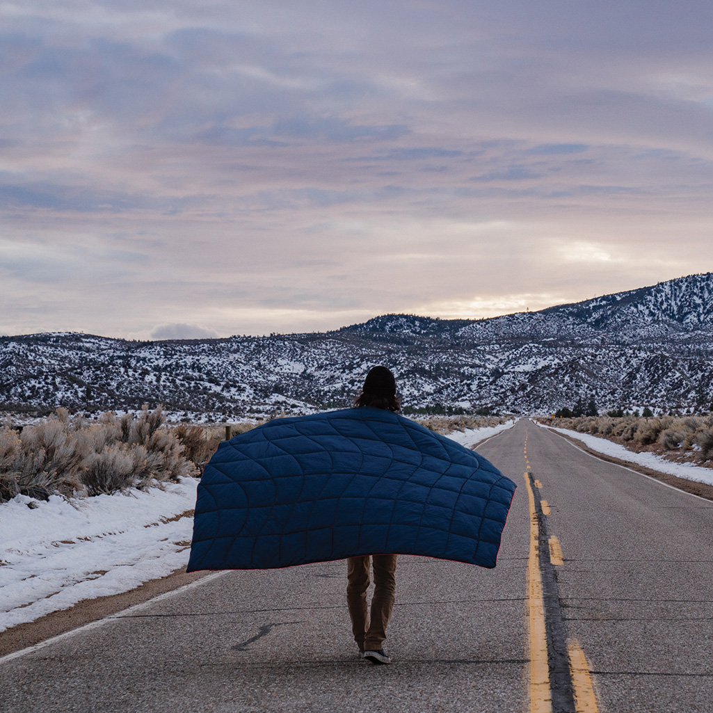 a person walking down a road holding a nano puffy blanket behind them with their arms stretched out 
