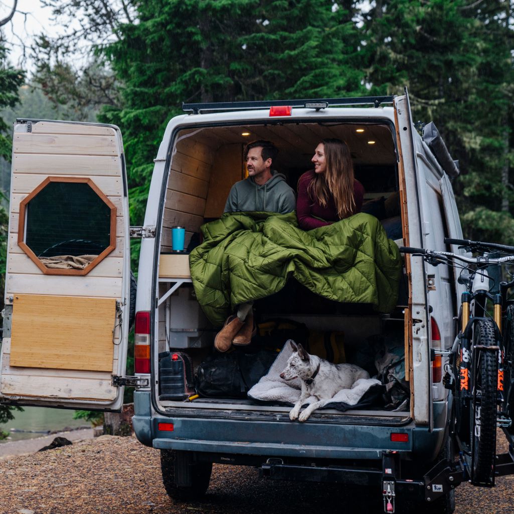 A couple sits comfortably in their camper van, wrapped in a cozy blanket, enjoying the scenic view outside.