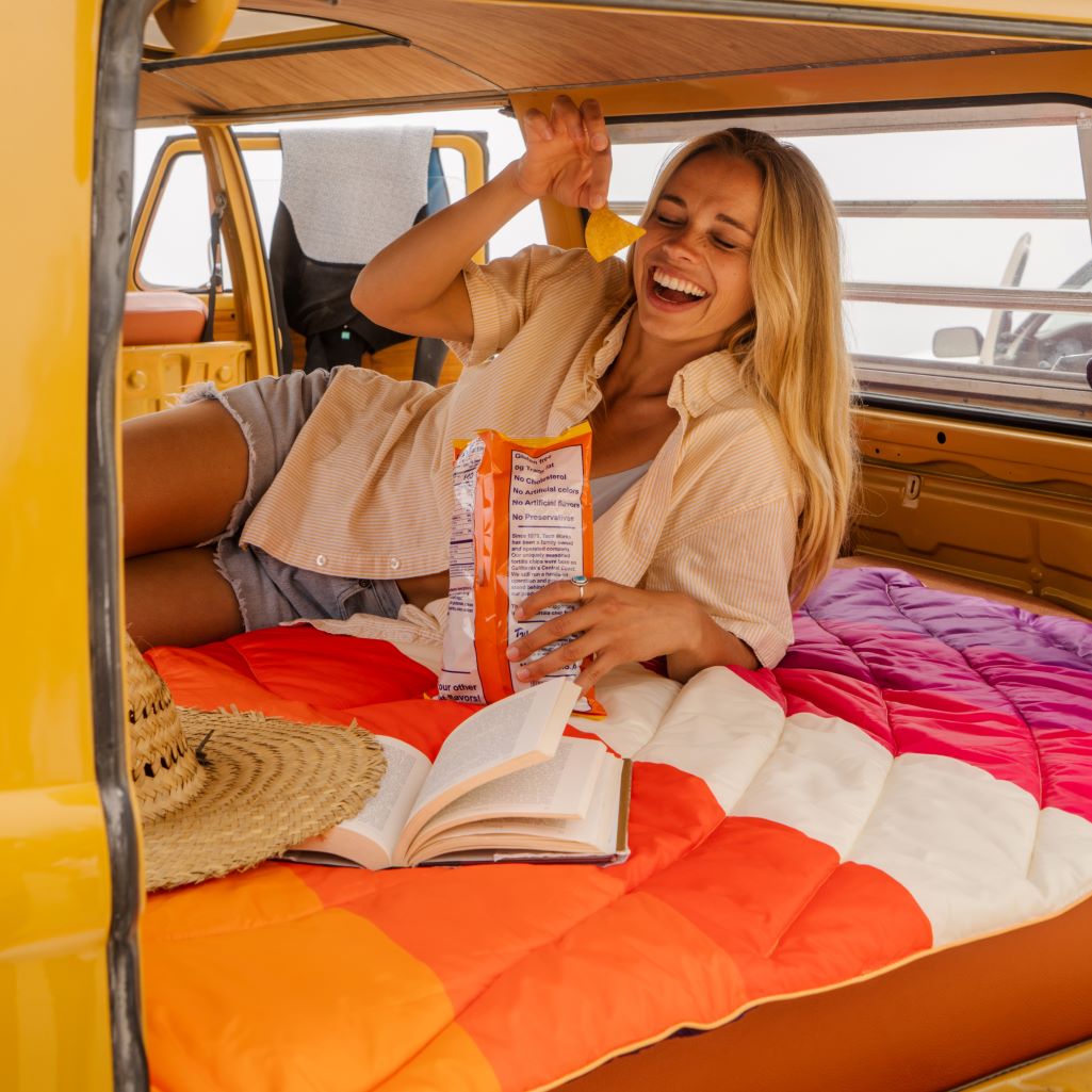 A girl chilling and looking worry-free on a Rumpl puffy blanket with a book and some snacks in the back of her camper van.
