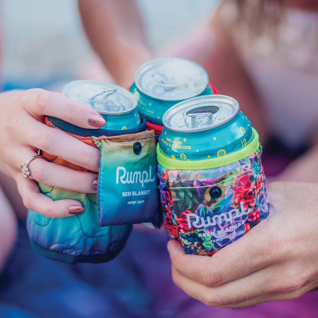 A close up of three people's beers are they share a 'Cheers!' moment. The cans all sport a Rumpl Beer Blanket in different prints.