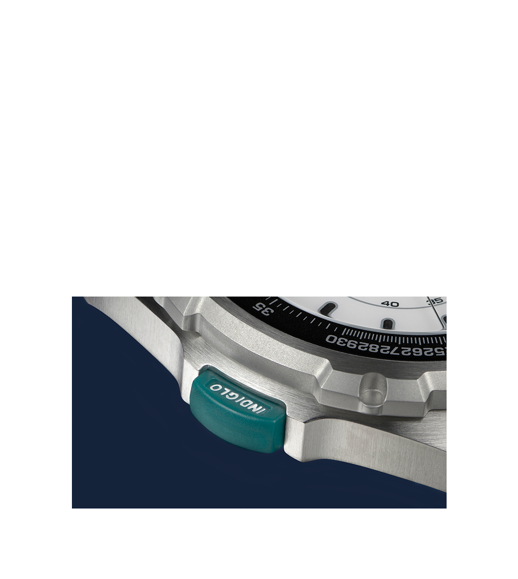 A detailed macro shot of the Timex 1995 Intrepid Reissue watch, highlighting the teal-colored INDIGLO button on the brushed stainless steel case. The button, engraved with 'INDIGLO' in white text, is set against the black rotating bezel and crisp white dial, emphasizing the watch's signature backlight feature.