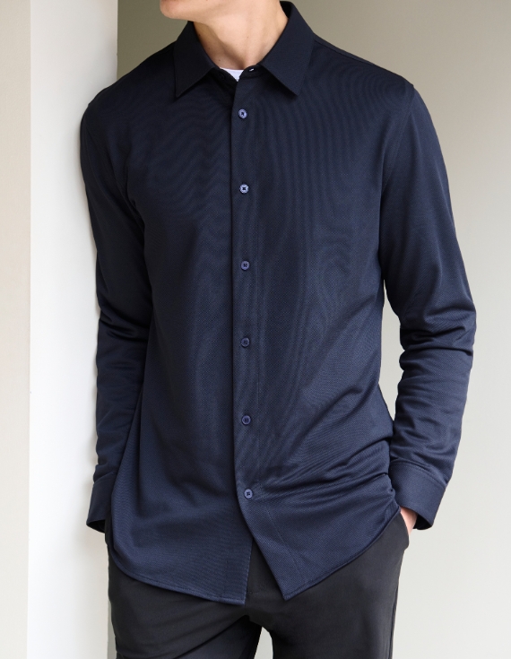 Man wearing a navy long-sleeve button-up shirt with a subtle textured pattern, paired with black trousers, standing casually against a light-colored wall