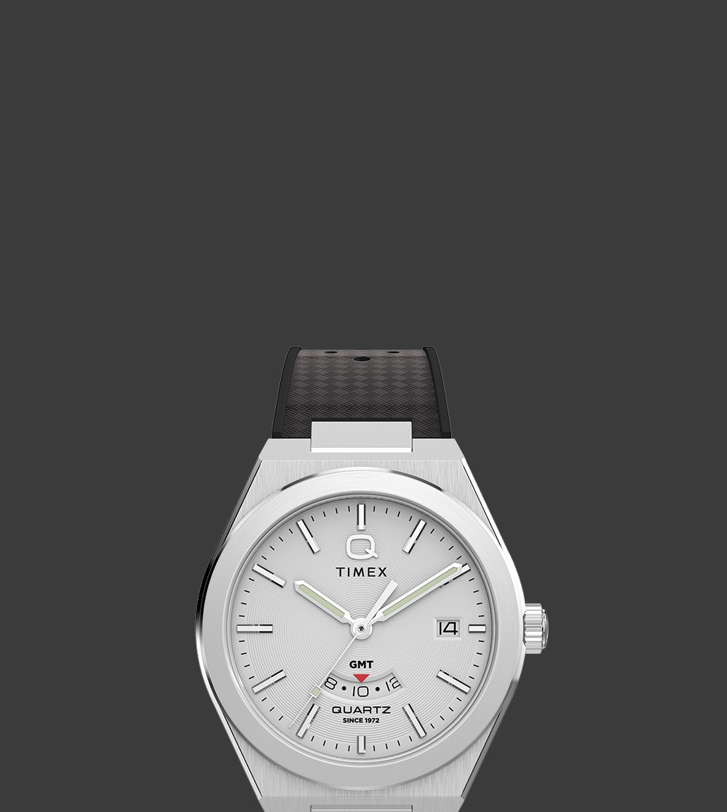 Front view of the Q Timex® Continental GMT watch with a silver-tone case and dial, black hour markers and hands, black strap with textured detail, and a GMT sub-dial located above 6 o’clock.