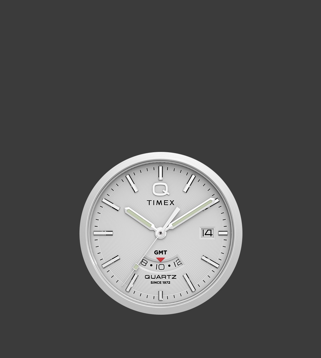 Close-up of the Q Timex® Continental GMT watch dial showcasing concentric dial texture, GMT sub-dial with red arrow hand, and polished hour markers against a silver background.