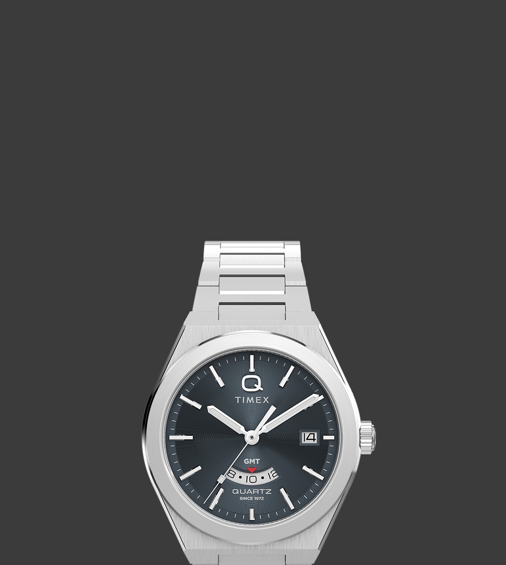 Front-facing view of the Q Timex® Continental GMT watch featuring a sleek brushed stainless steel case and bracelet, dark green textured dial, and red-accented GMT sub-dial above 6 o'clock.