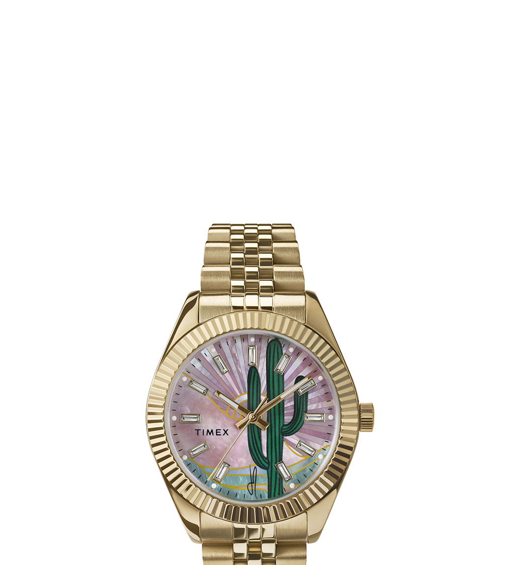 Front-facing product image of the Timex x Jacquie Aiche gold-tone watch. The dial features a hand-drawn green cactus with a radiant mother-of-pearl pink background, gold-tone markers and hands, and a five-link bracelet with a fluted bezel.