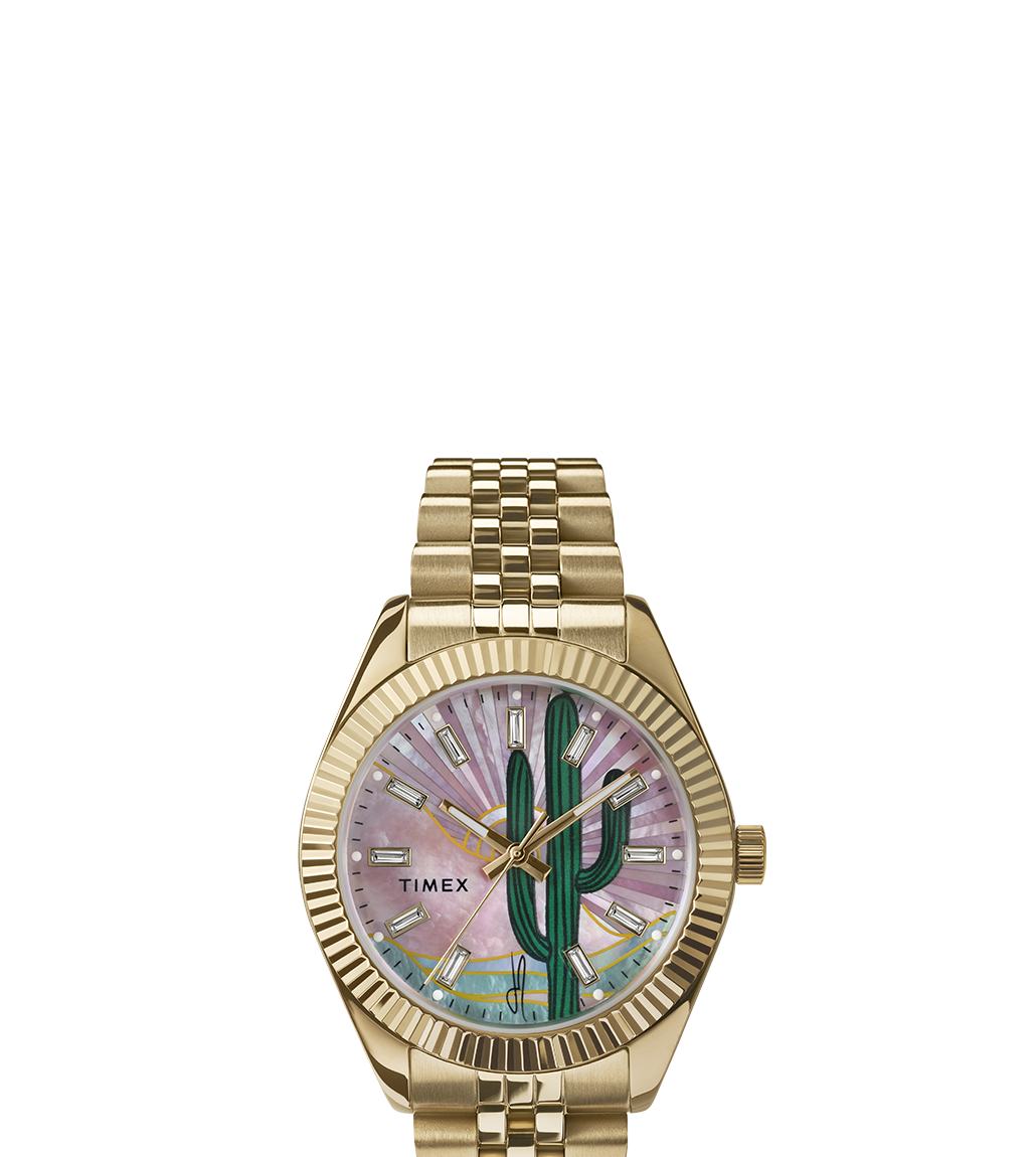 Front-facing product image of the Timex x Jacquie Aiche gold-tone watch. The dial features a hand-drawn green cactus with a radiant mother-of-pearl pink background, gold-tone markers and hands, and a five-link bracelet with a fluted bezel.