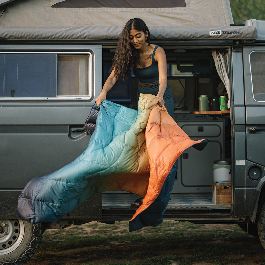 a woman standing on a camper van, dusting out her Rumpl NanoLoft® Puffy Blankets