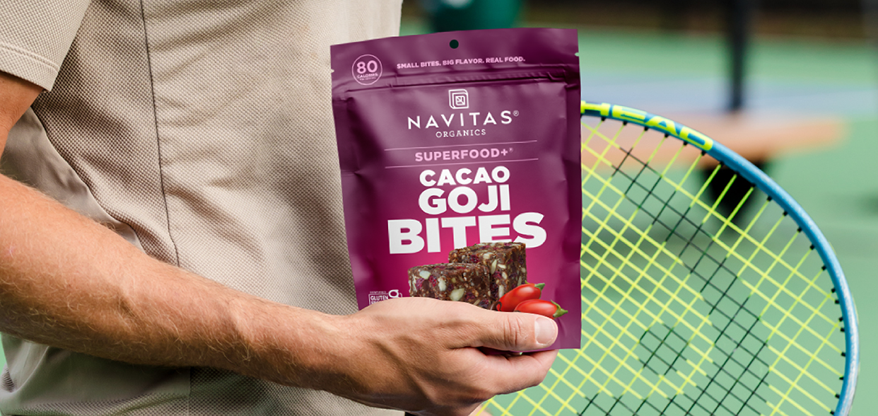 A person holding a tennis racket in one hand and a bag of Navitas Organics Superfood+ Cacao Goji BITES in the other, standing on a tennis court.