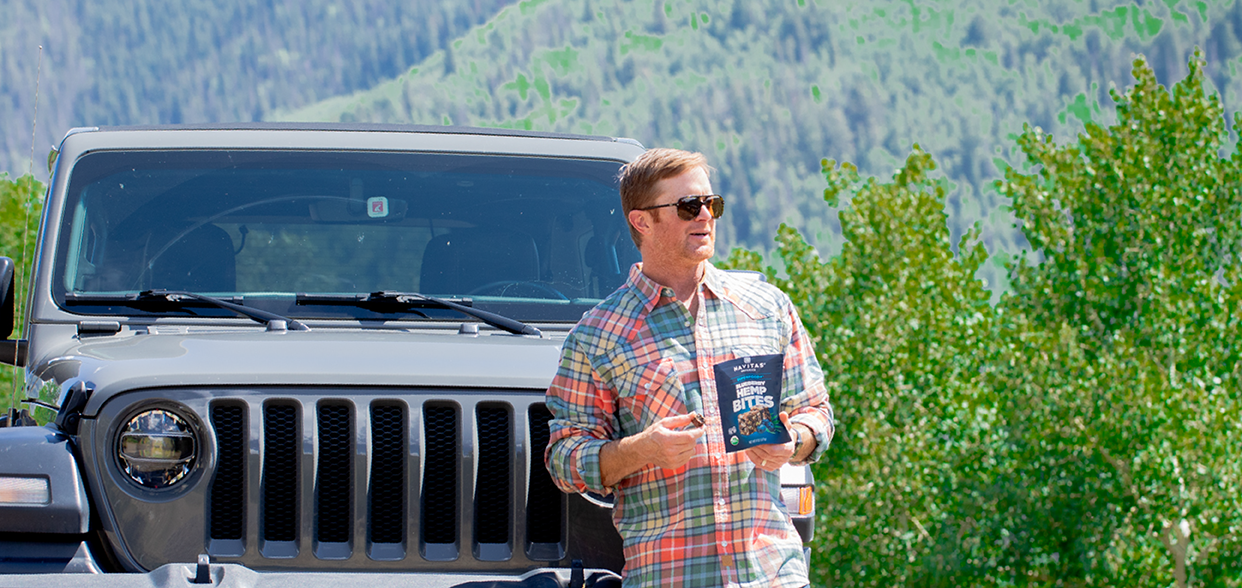 Person holding a bag of Navitas Organics Superfood+ Blueberry Hemp BITES standing in front of a Jeep in a mountainous setting.