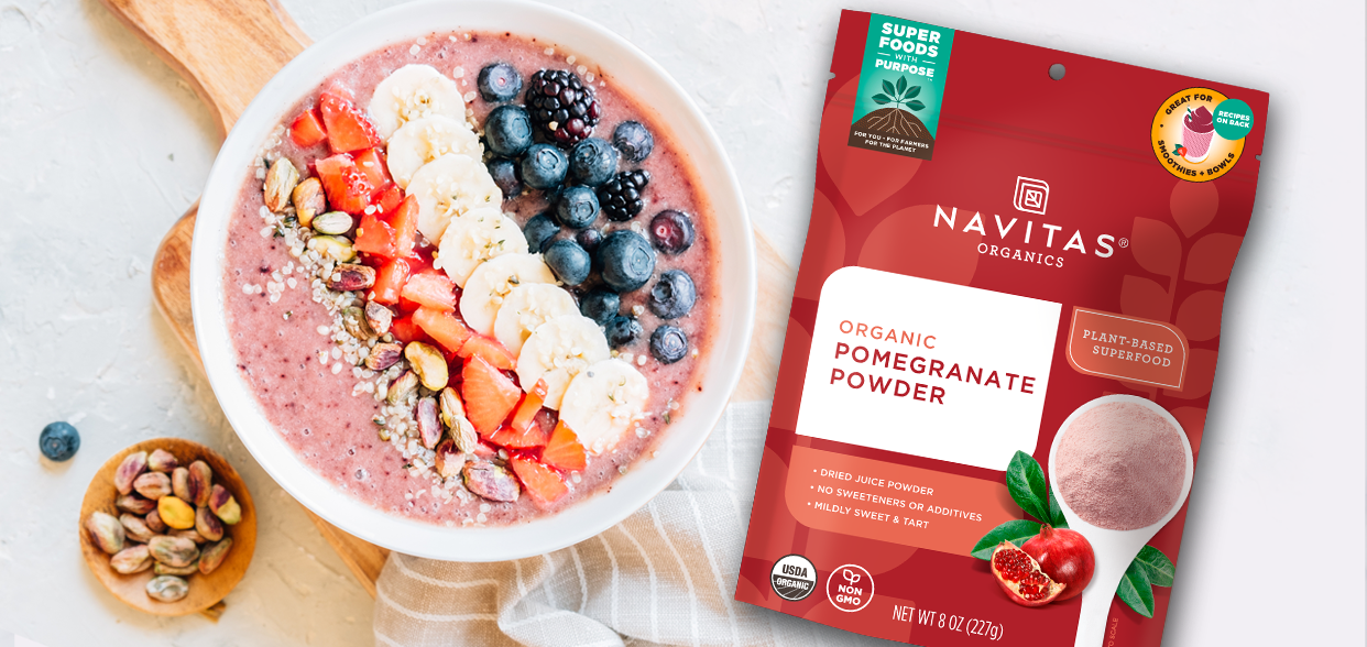 A bowl of fruit smoothie topped with sliced bananas, strawberries, blueberries, and mixed seeds sits next to a package of Navitas Organics Organic Pomegranate Powder.