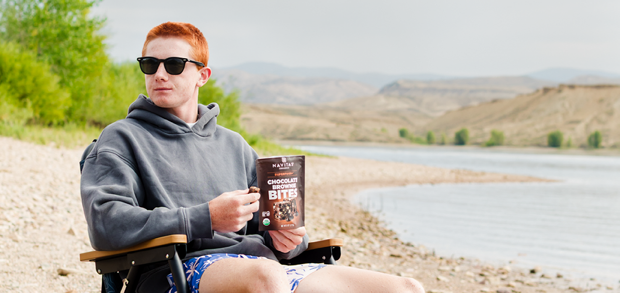 Person sitting in a camping chair by a river, holding a bag of Navitas Organics Superfood+ Chocolate Brownie BITES, with hills in the background.