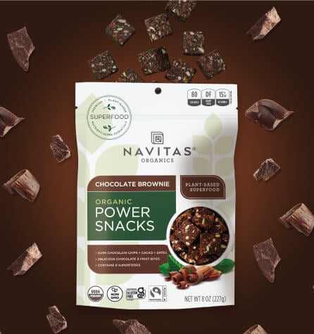 A package of Navitas Organics Chocolate Brownie Power Snacks surrounded by chocolate chunks against a brown background.