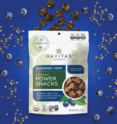A package of Navitas Organics Blueberry Hemp Power Snacks surrounded by blueberries and hemp seeds against a blue background.