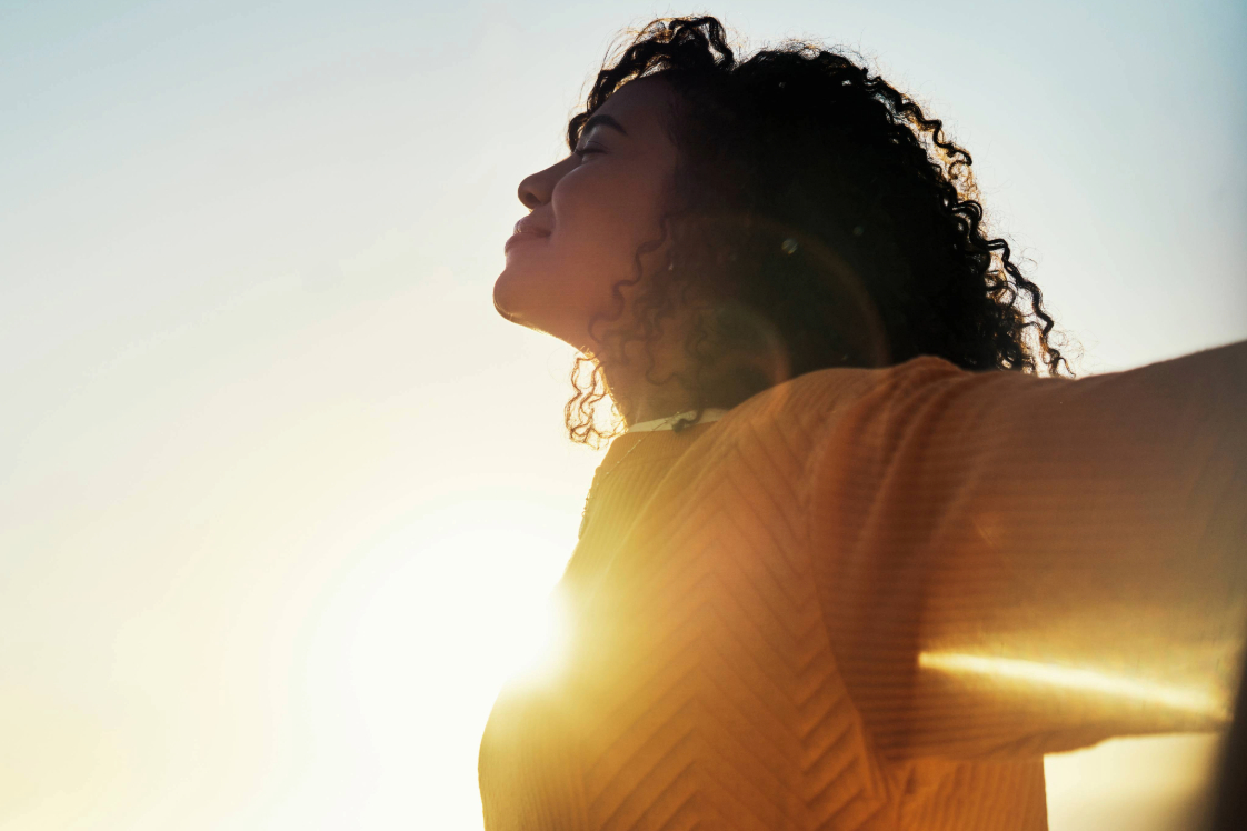Woman in yellow sweater outdoors opening her arms with the sun in the background.