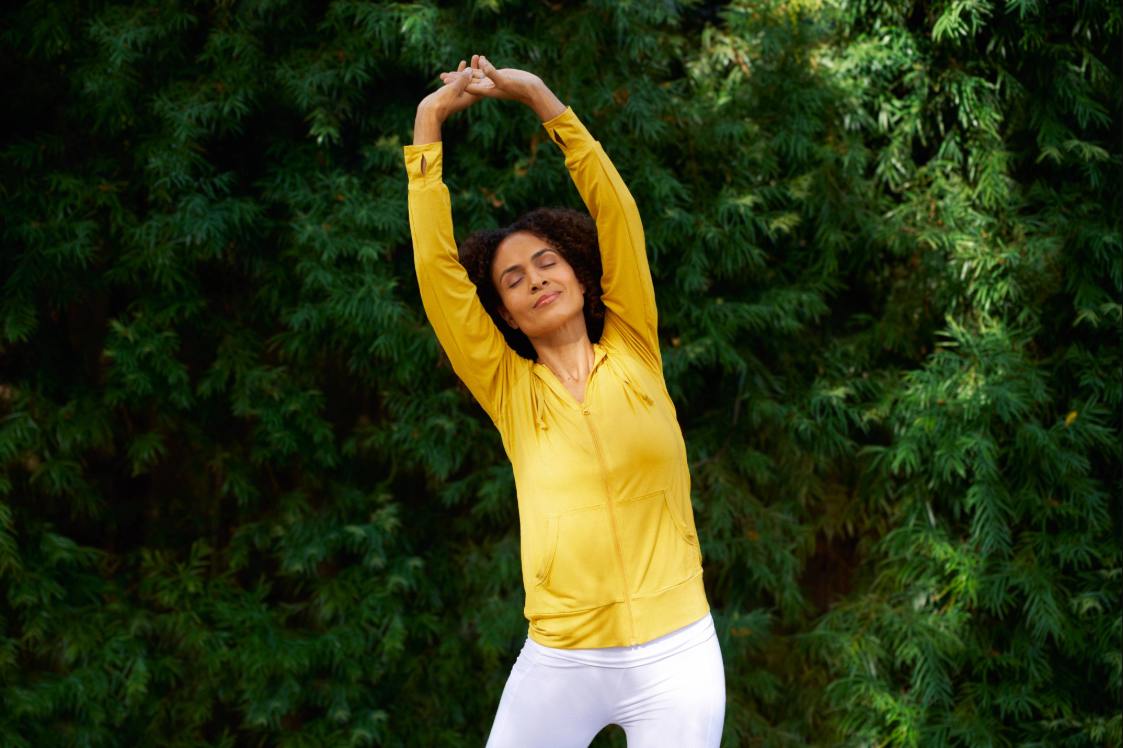 Person stretching arms overhead outdoors with green trees behind