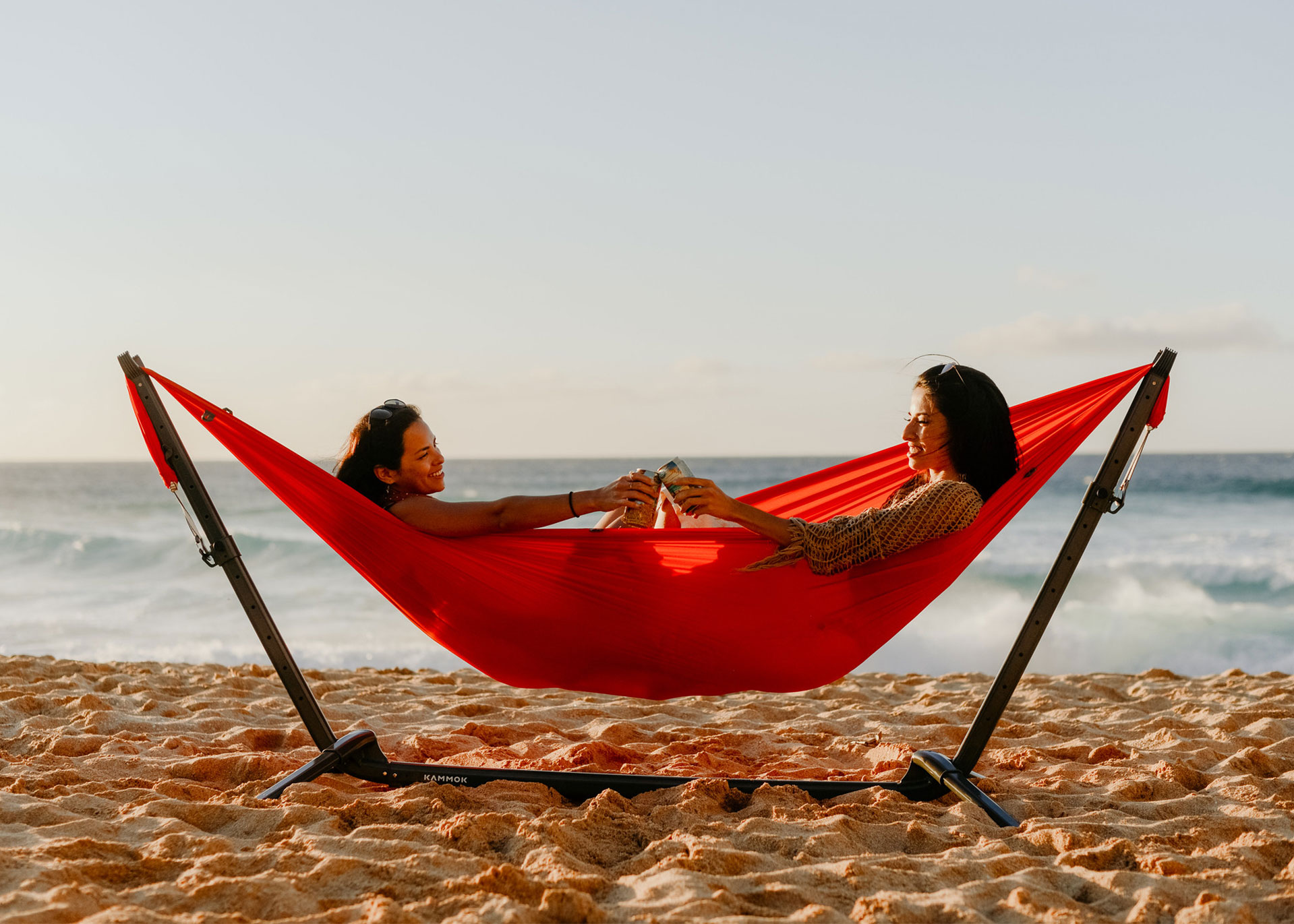 Two women sitting in a Kammok Roo Double camping hammock made from 100% recycled fabric attached to a Swiftlet hammock stand on the beach.
