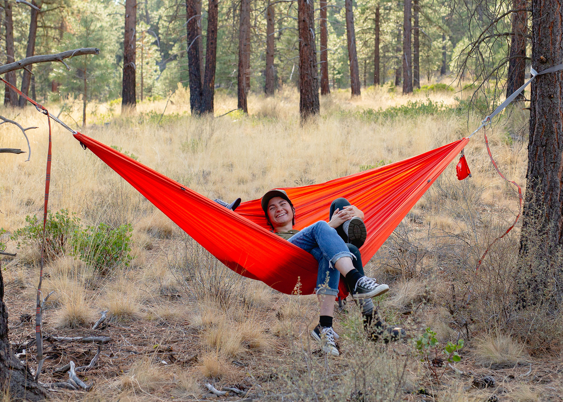 Two people sit in an Ember Oranger Roo Double Camping Hammock hung from trees in the woods.