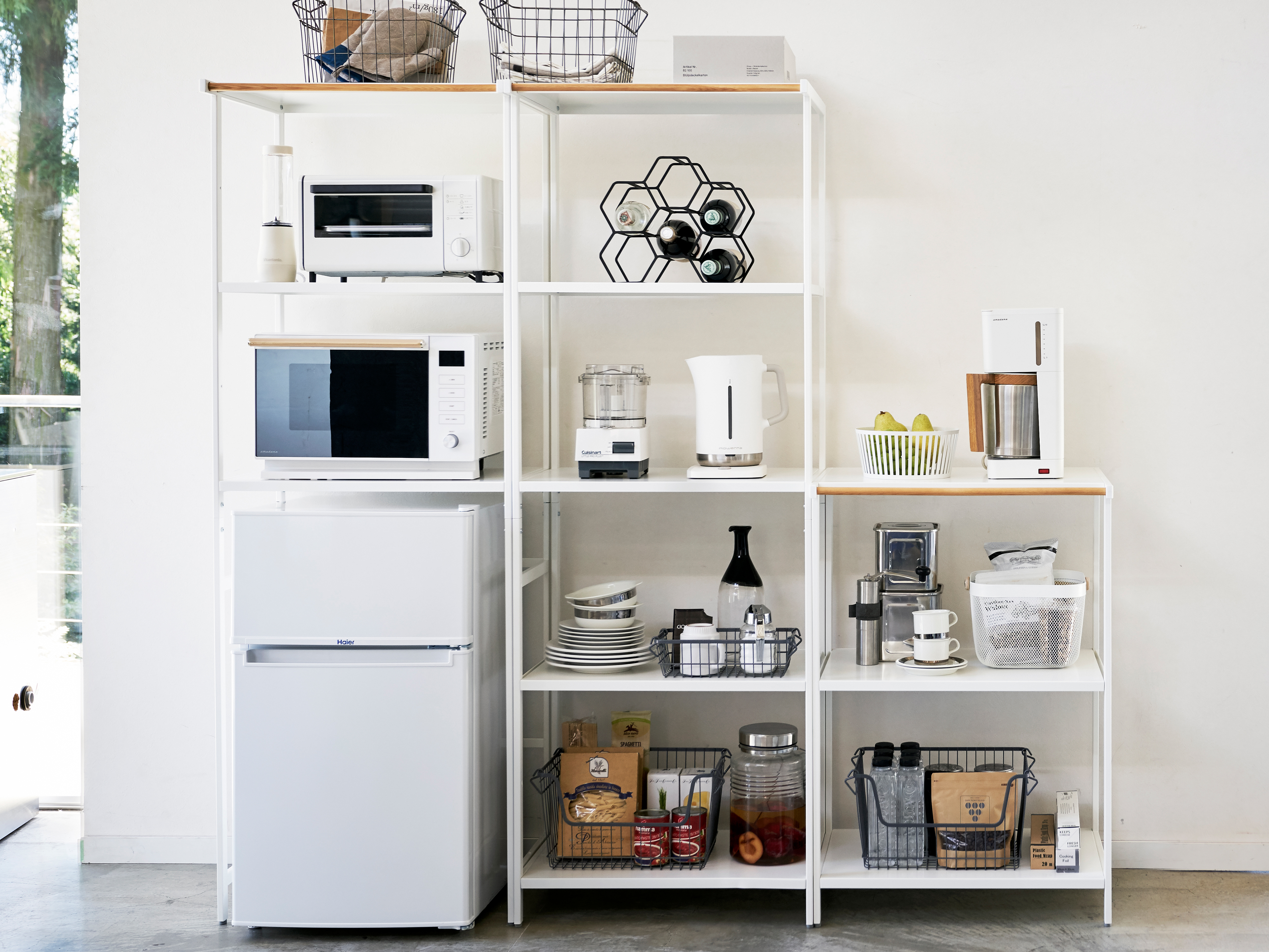 Three white minimalist shelving units with light wood tops stand against a white wall, filled with kitchen appliances, cookware, and pantry items in an organized display.