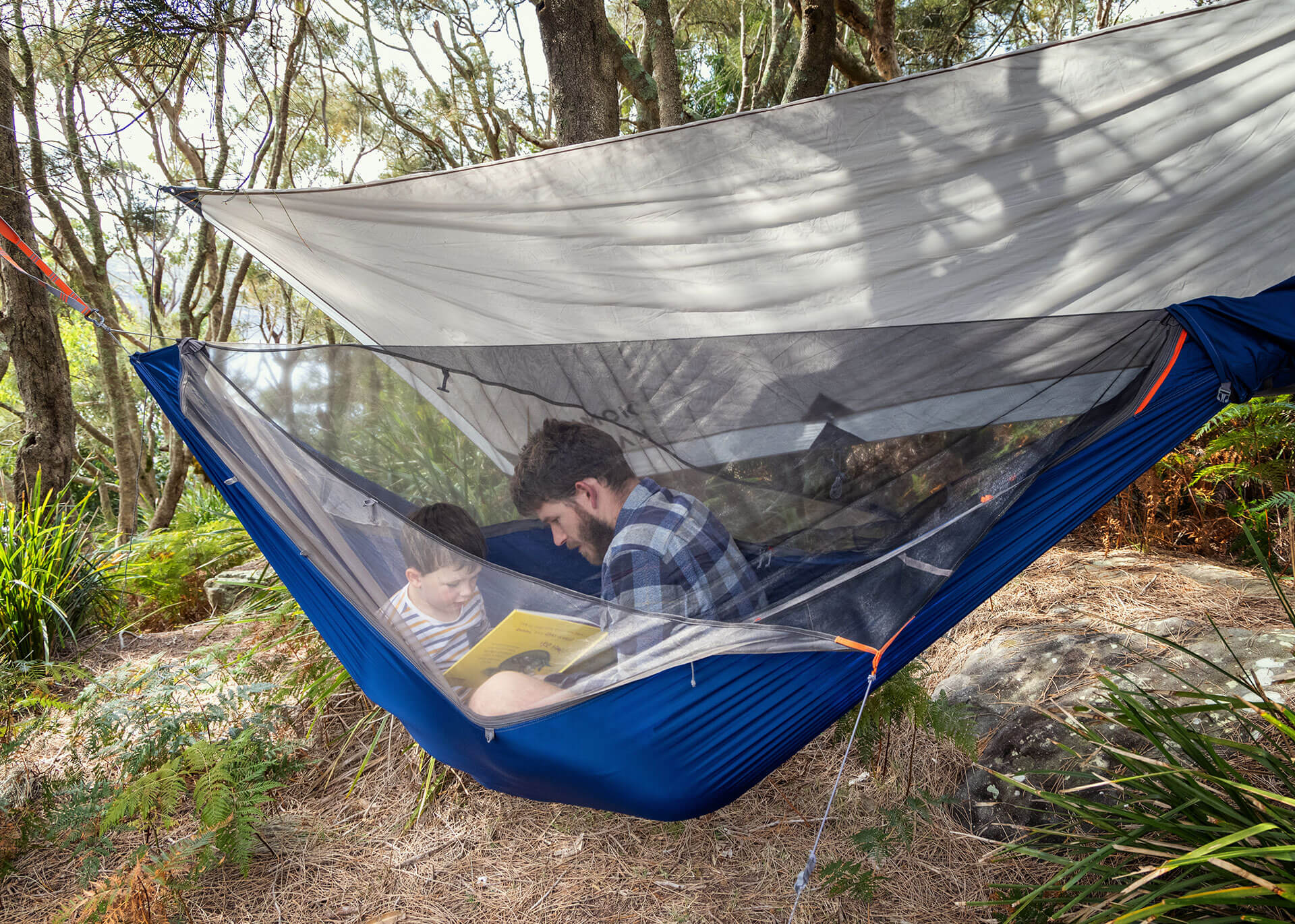 Man and child sitting in a Kammok Mantis all-in-one hammock tent with bug net and weather shelter hung between trees.