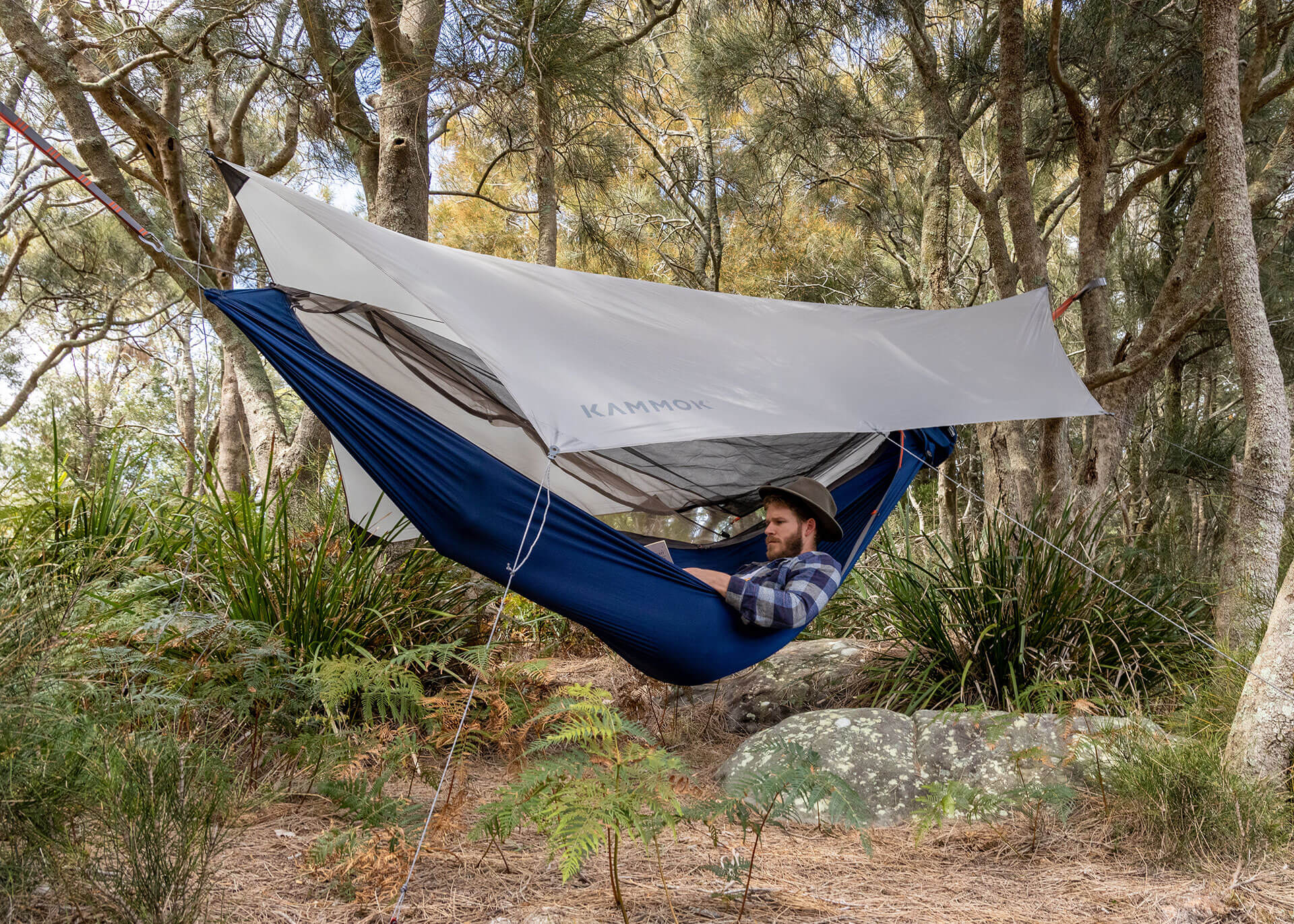 Man laying down Kammok Mantis all-in-one hammock tent with bug net, weather shelter, hammock straps, and hammock, hung between trees.