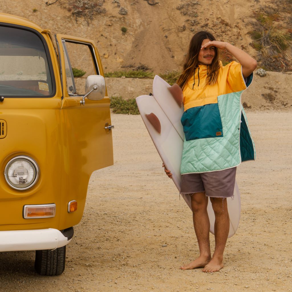 Surfer wearing a colorful Rumpl puffy poncho, holding a surfboard, standing outside a yellow van on the beach