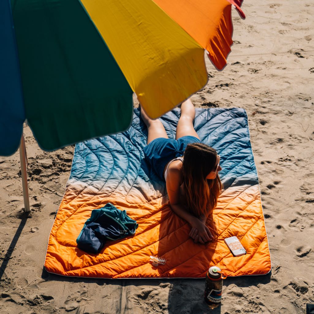 A girl lying on a vibrant Rumpl Original Puffy Blanket on a sandy beach.