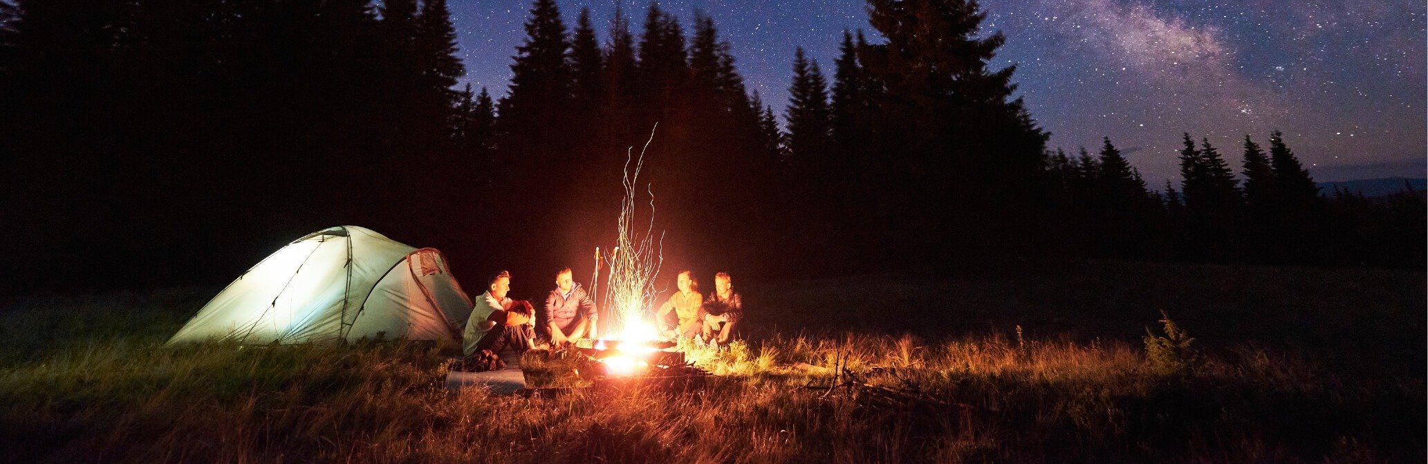 Friends camping in the forest against a starry night. Group is in front of a campfire next to a large tent.