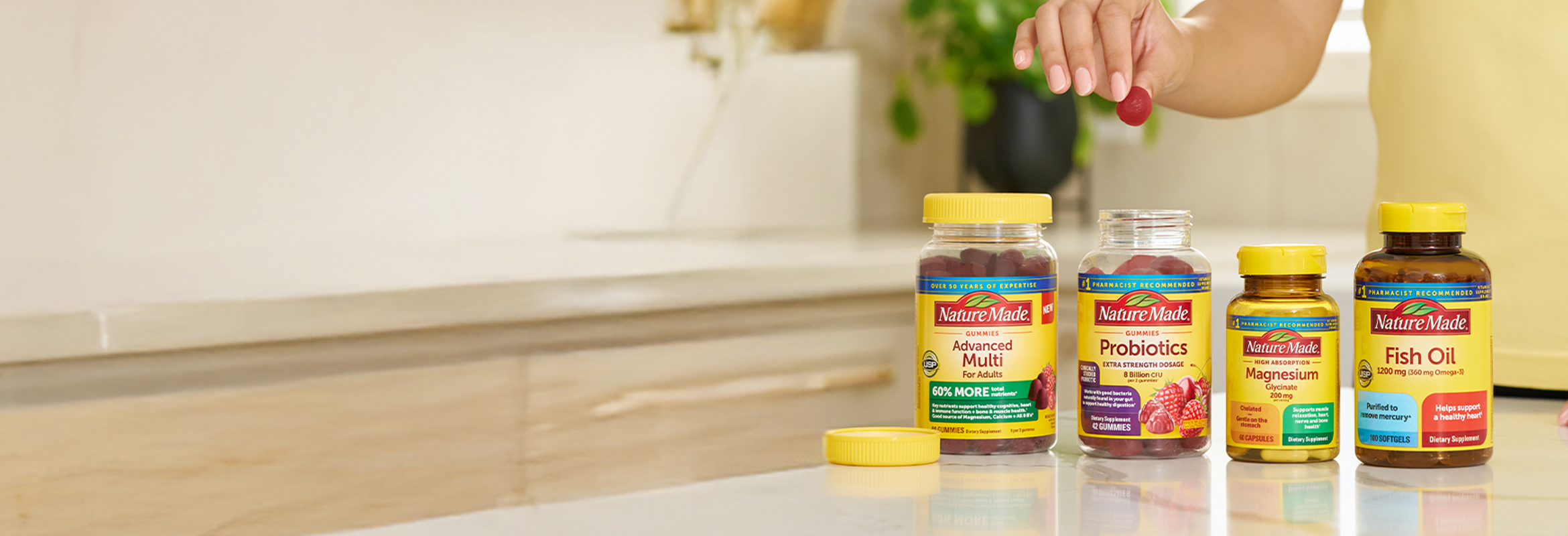 Hand picking a gummy from Nature Made supplement bottles on a kitchen counter, featuring Advanced Multi, Probiotics, Magnesium, and Fish Oil.