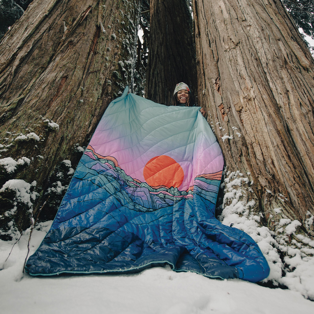 Artist, Brooklyn Bell holds up the Original Puffy Blanket - Skyline Divide for us to see. The unique print blanket looks vibrant against a surrounding snowy landscape.