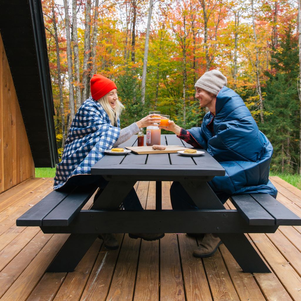 Two people sitting at a picnic table, wrapped in warm puffy blankets and enjoying a meal outdoors on a chilly day, surrounded by nature.