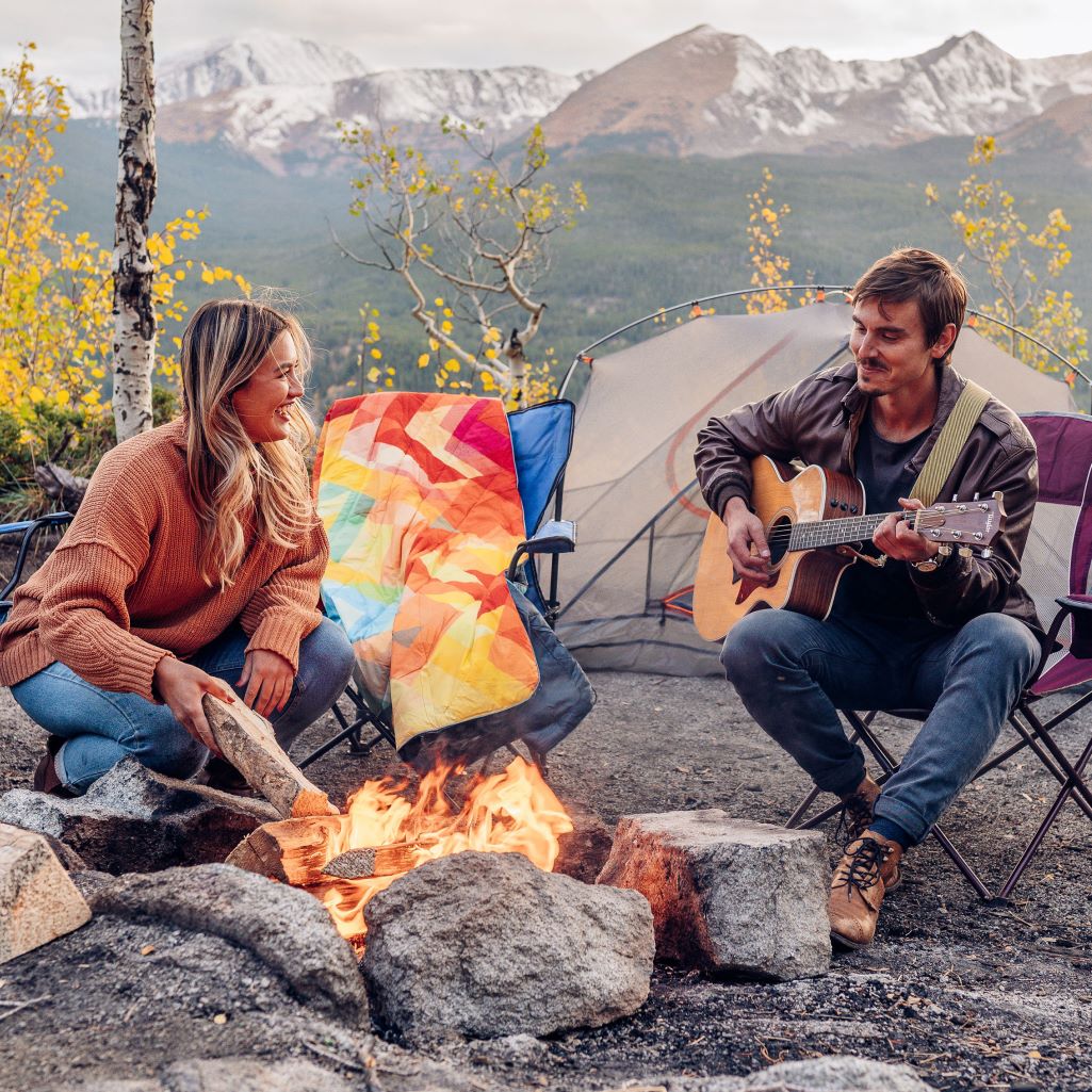 Two friends having a relaxing time at camp, by the campfire with a few Rumpl blankets for extar coziness.