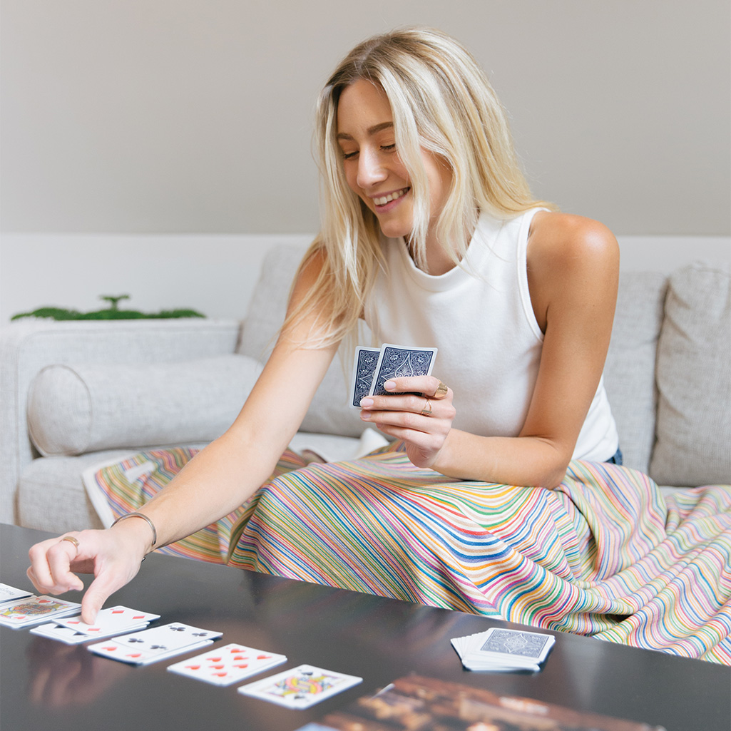 A woman plays cards on her couch while cozying up with the Rumpl CozyHemp™ cotton and hemp blend natural fiber blanket.