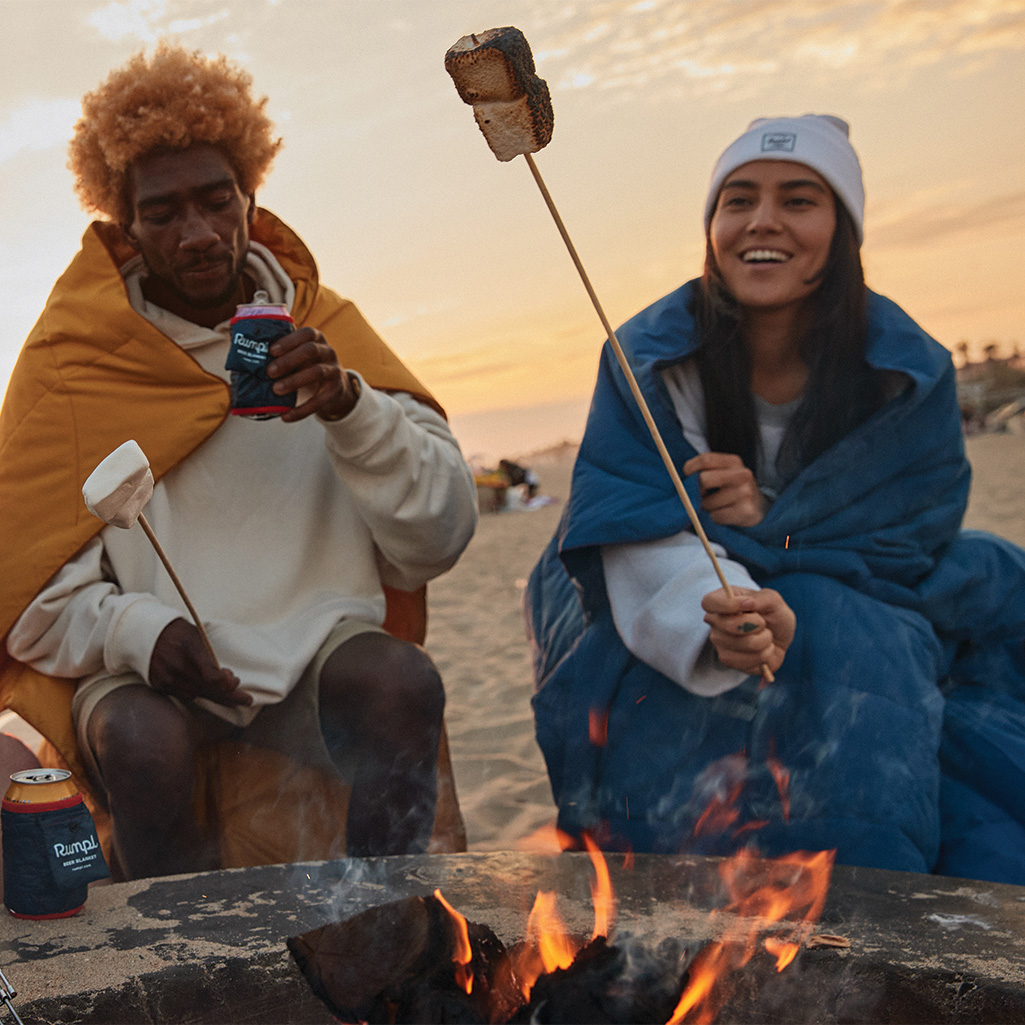 a pair of friends roasting marshmallows on a stick over a campfire while wearing the NanoLoft® Flame-resistant blanket over their shoulders with the Cape Clips®️