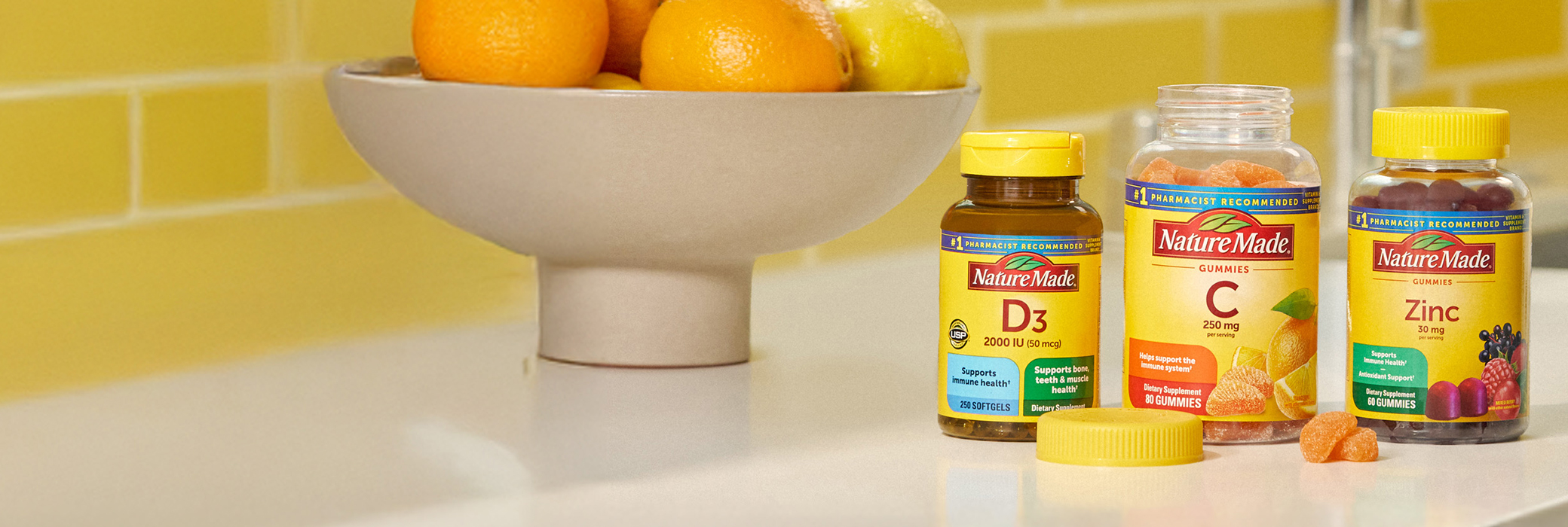 Nature Made Vitamin D3, Vitamin C and Zinc supplements on a kitchen counter top with a bright yellow tile background.