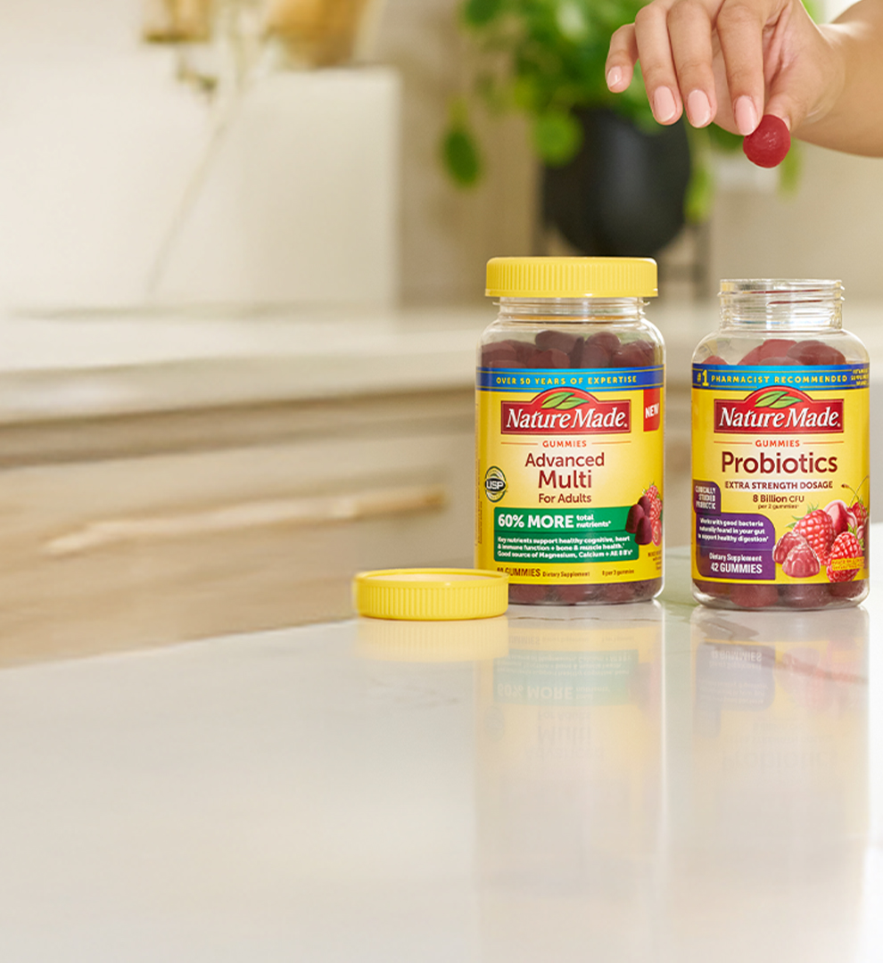 Hand picking a gummy from Nature Made supplement bottles on a kitchen counter, featuring Advanced Multi, Probiotics, Magnesium, and Fish Oil.