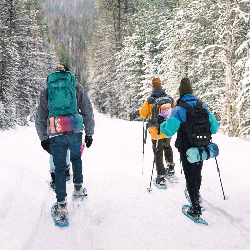 Three skiers walking on a snowy slope with vibrant-colored Rumpl blankets strapped to their backpacks.