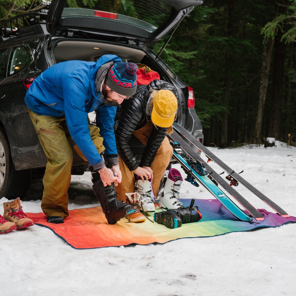 Two skiers using a Rumpl Everywhere Mat as a snowproof layer next to their car as they adjust their ski gear.