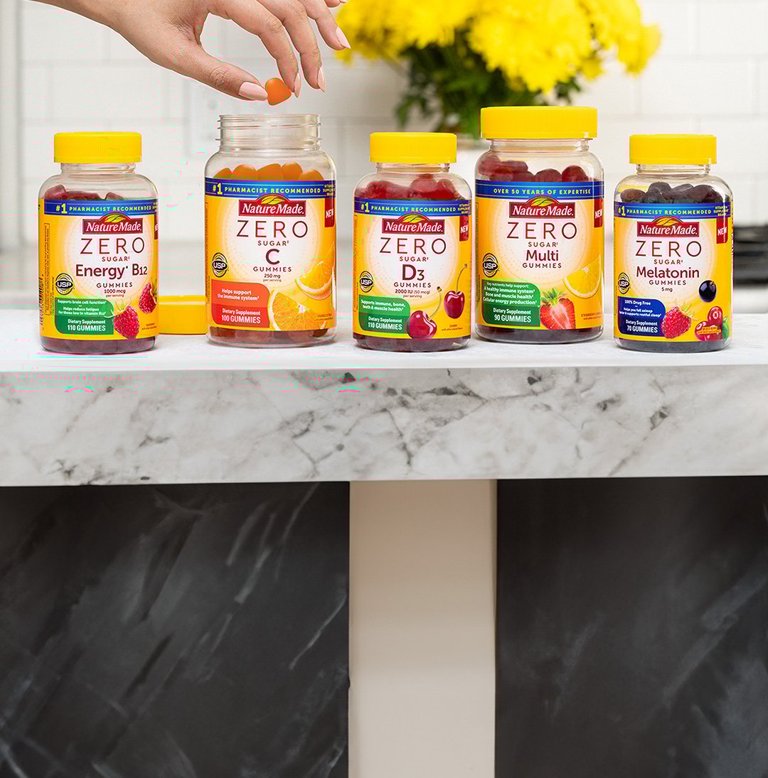 Person reaching for a gummy from an open bottle of Nature Made Zero Sugar vitamins on a kitchen counter.