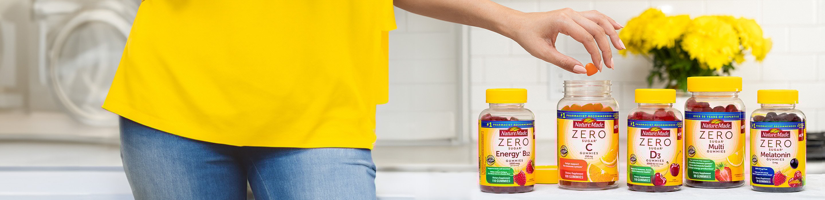 Person in a yellow top reaching for a gummy from an open bottle of Nature Made Zero Sugar vitamins on a kitchen counter.