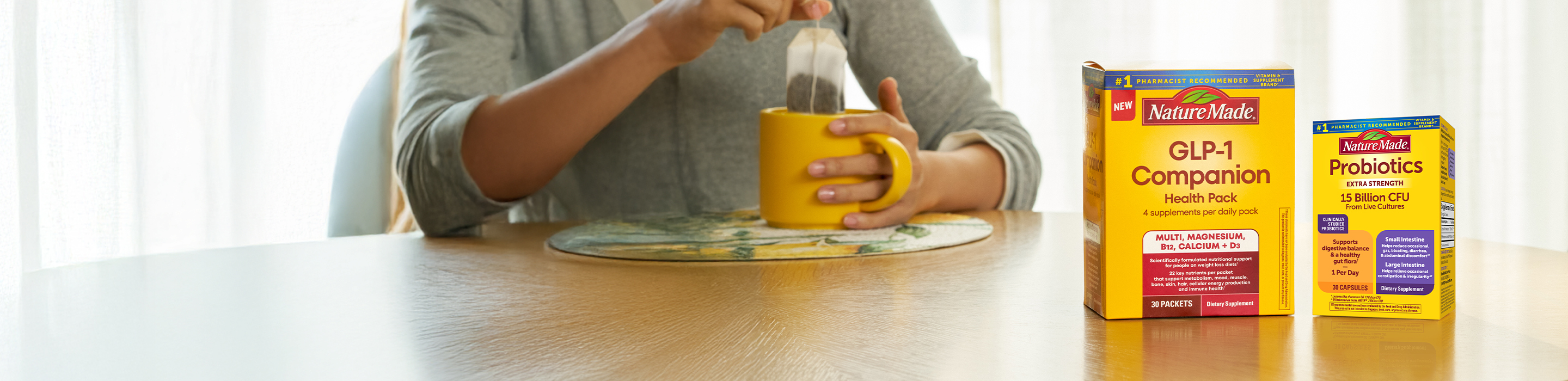 Nature Made GLP-1 Companion Health Pack and Probiotics Extra Strength boxes on a wooden table next to a person preparing tea in a yellow mug.