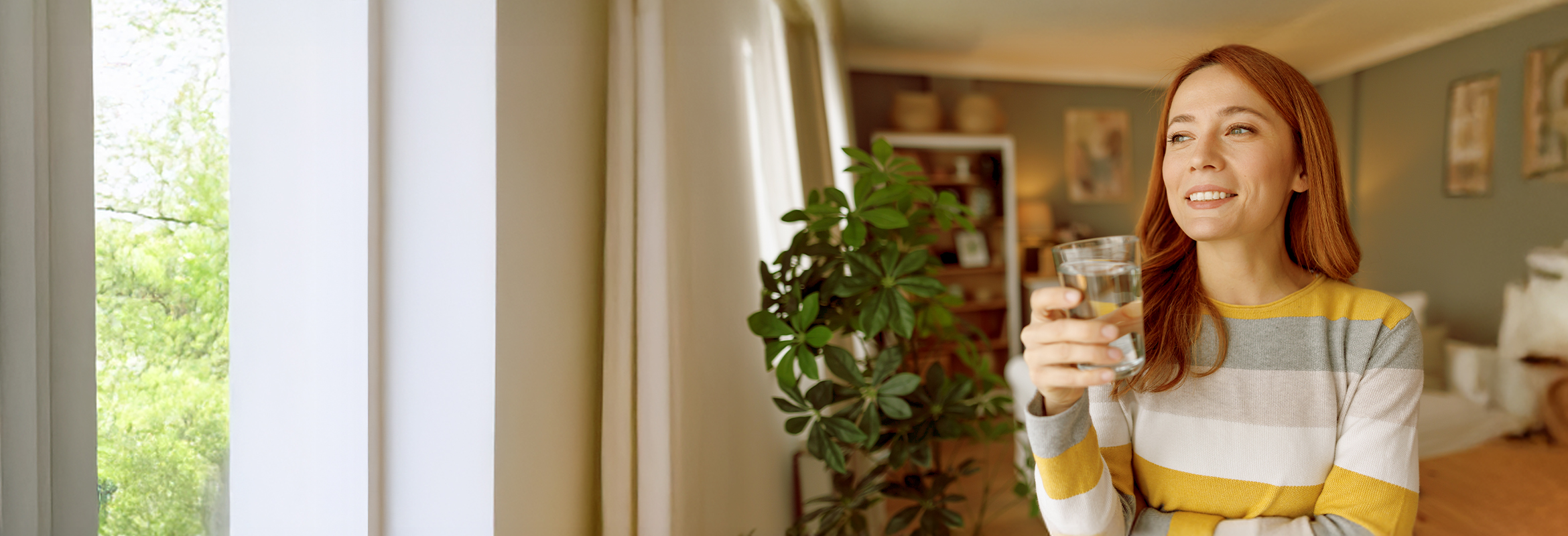 Person in a yellow, grey and white stripped sweater standing next to a plant in a living room with a glass of water in her hand.