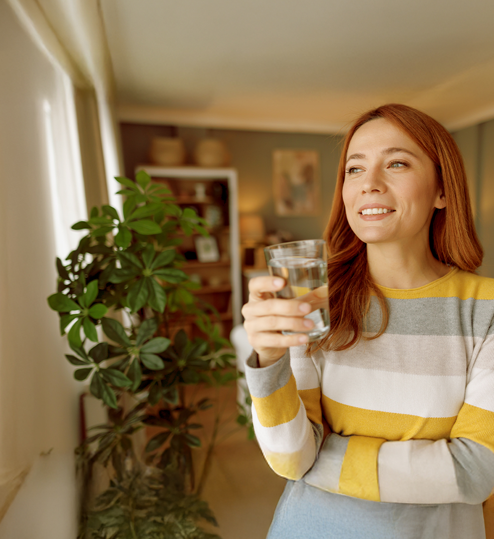 Person in a yellow, grey and white stripped sweater standing next to a plant in a living room with a glass of water in her hand.