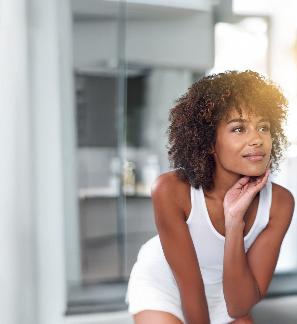 Woman with curly hair in white top looking in the mirror and touching her face.