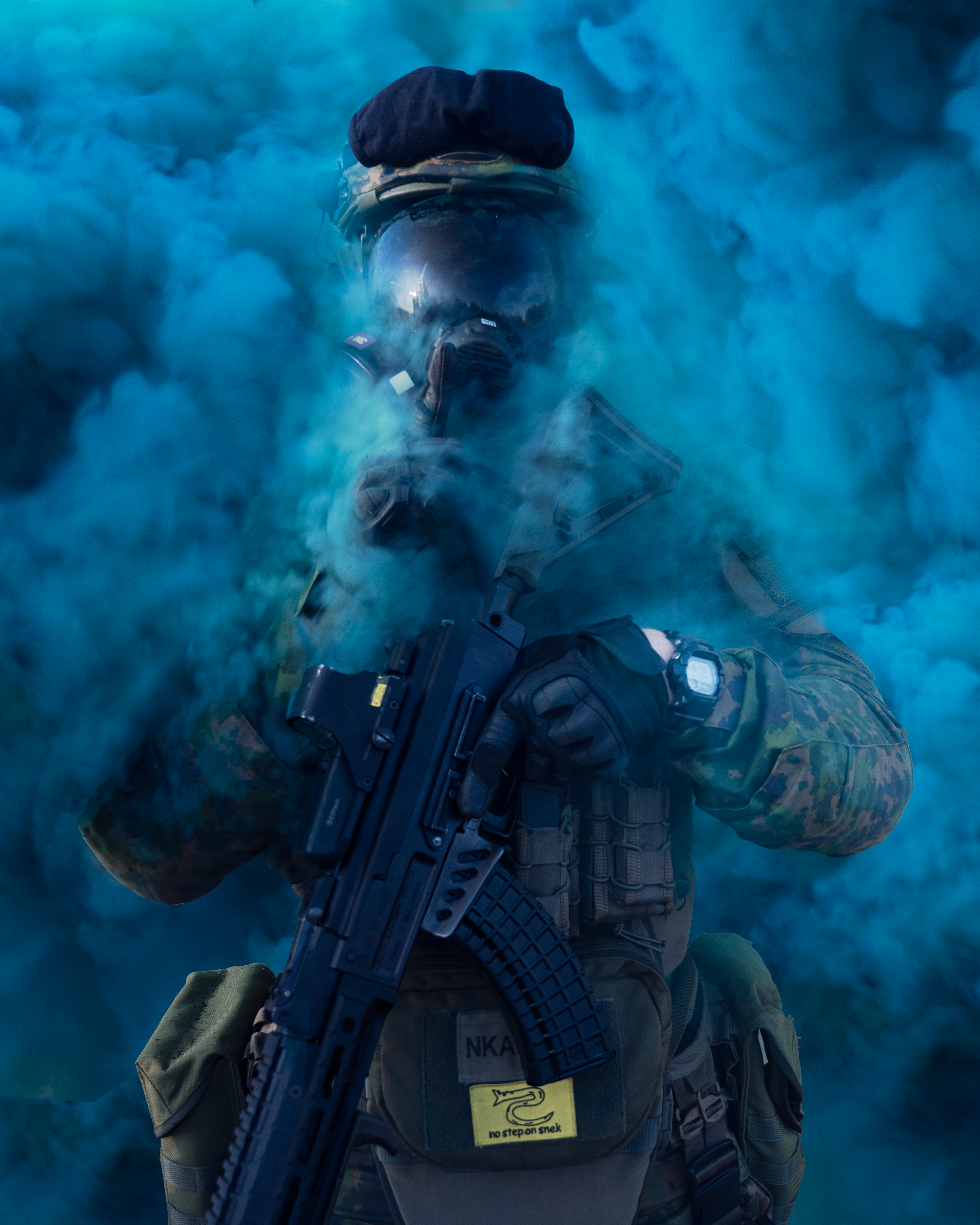 Man standing in blue smoke with a rifle in hand. He has a gas mask.